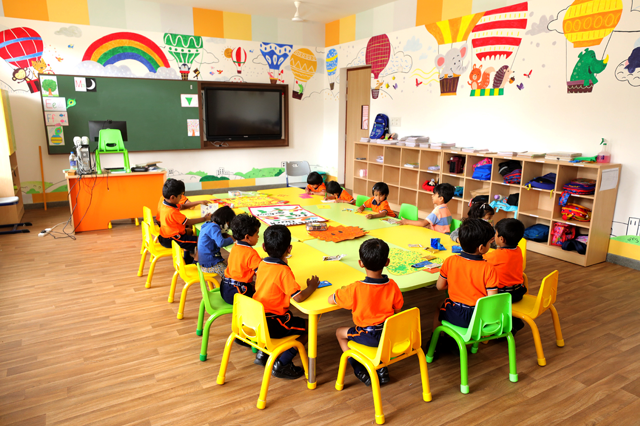 Colorful kindergarten classroom with children in orange uniforms seated around a yellow table doing crafts.