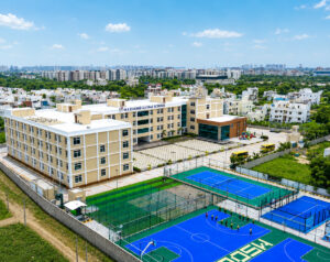 Aerial view of a school campus with beige buildings, blue sports courts and green fields, surrounded by residential blocks and trees.