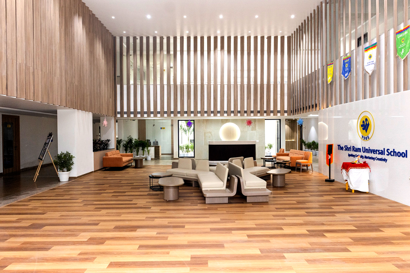 Spacious school lobby with wooden slat walls, a circular reception desk, beige sofas, and banners along the right wall showing the school name.